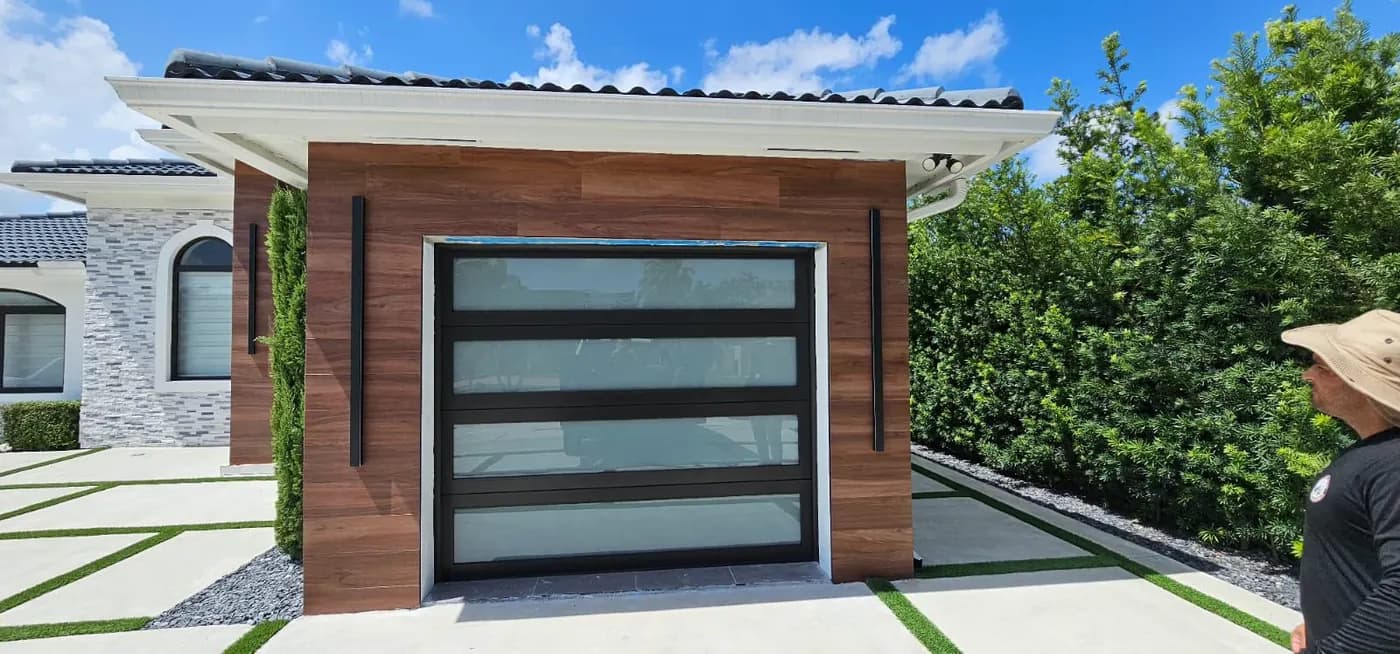 Black frame impact glass garage door on a modern wood-accent garage with turf-lined driveway